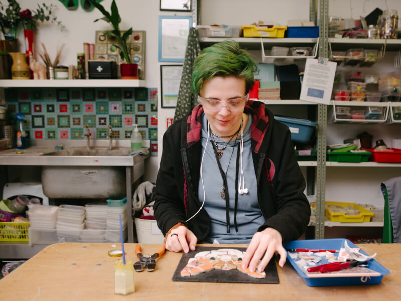 A person is sitting at a table creating a mosaic design of a cat's face using coloured tiles